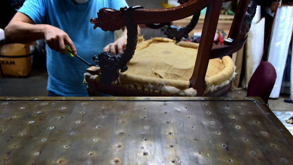 Craftsman working on a carved wood chair stripped to its burlap seat foundation in the upholstery shop