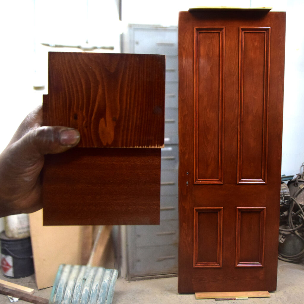 Hand holding a finished wood stain sample next to a refinished four-panel door in the shop