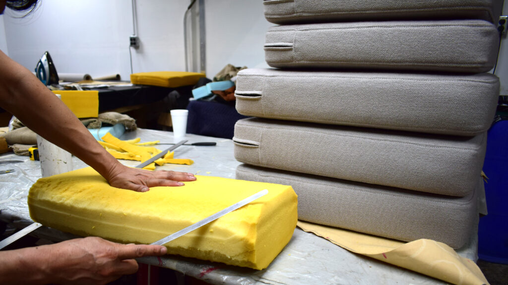 Upholsterer measuring and cutting a foam cushion core beside stacked finished cushions